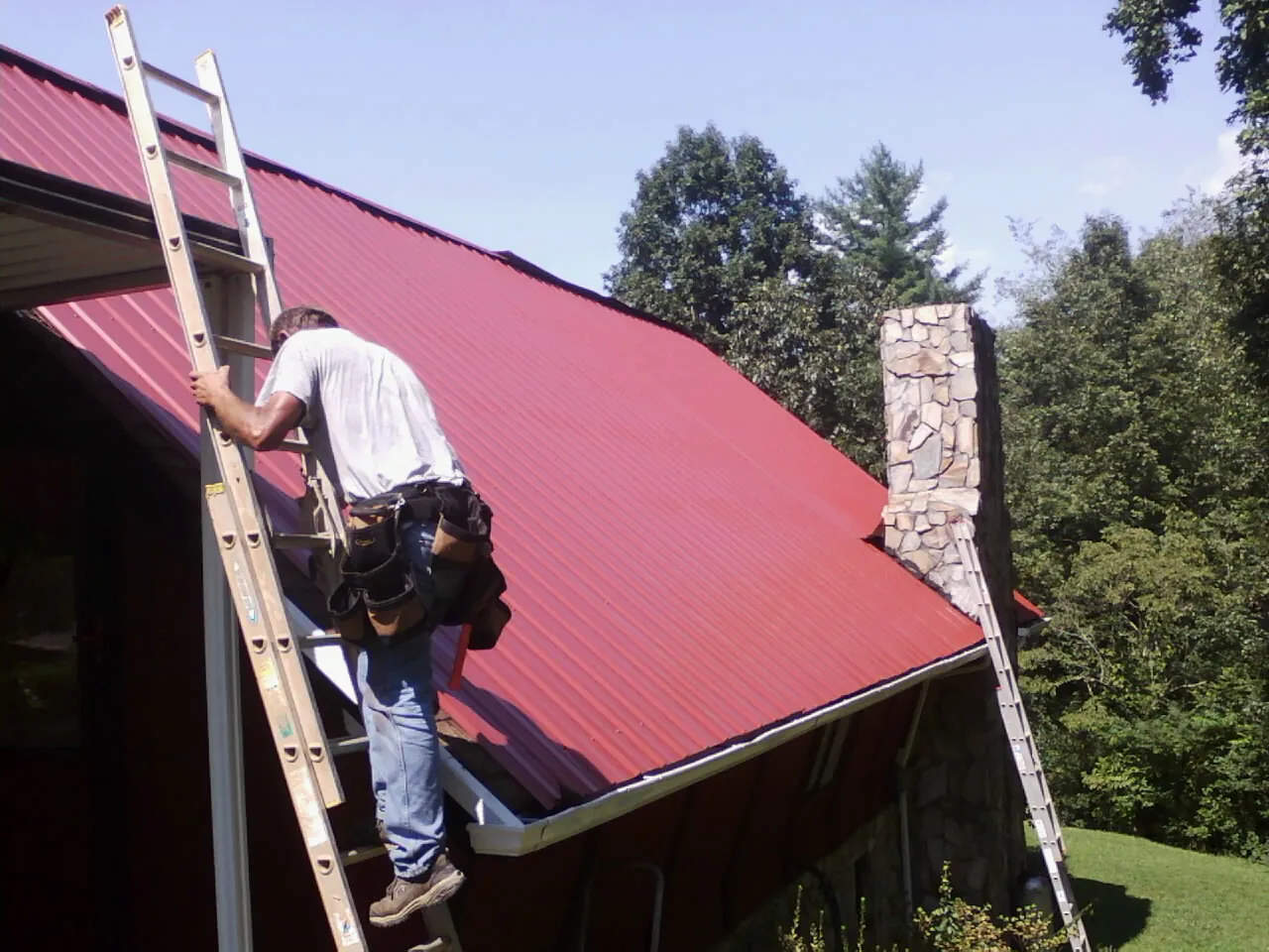 Roofer on a ladder beside a red metal roof and stone chimney — Soffit & Fascia Repair by Roofing Brothers in Rockbridge Baths